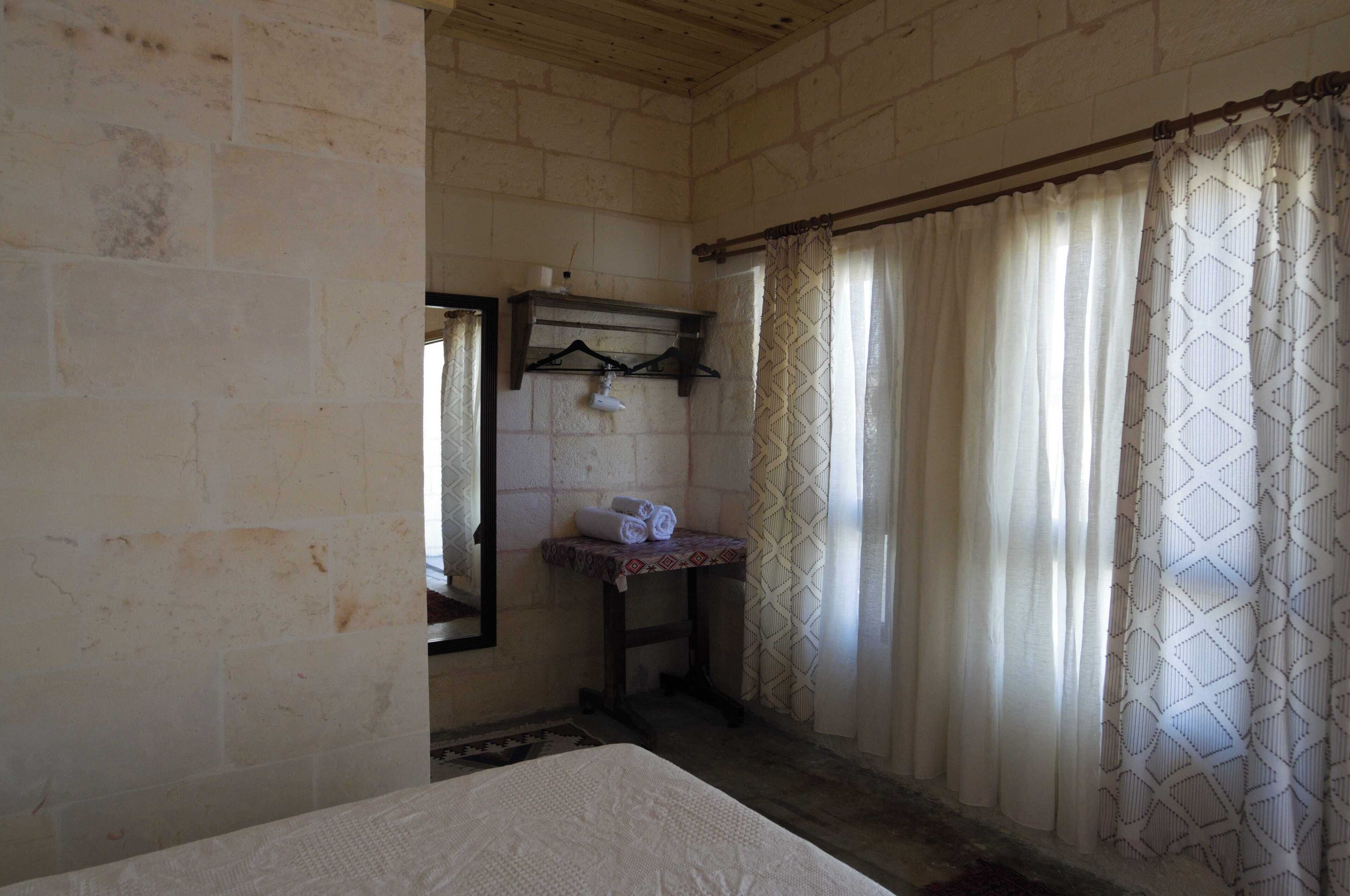 A small, dimly lit room with a bed, a small table with a towel, and a window with patterned curtains.