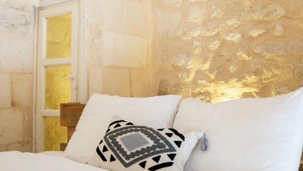 A bedroom with a stone wall, a wooden door, and a bed with white bedding and patterned pillows.