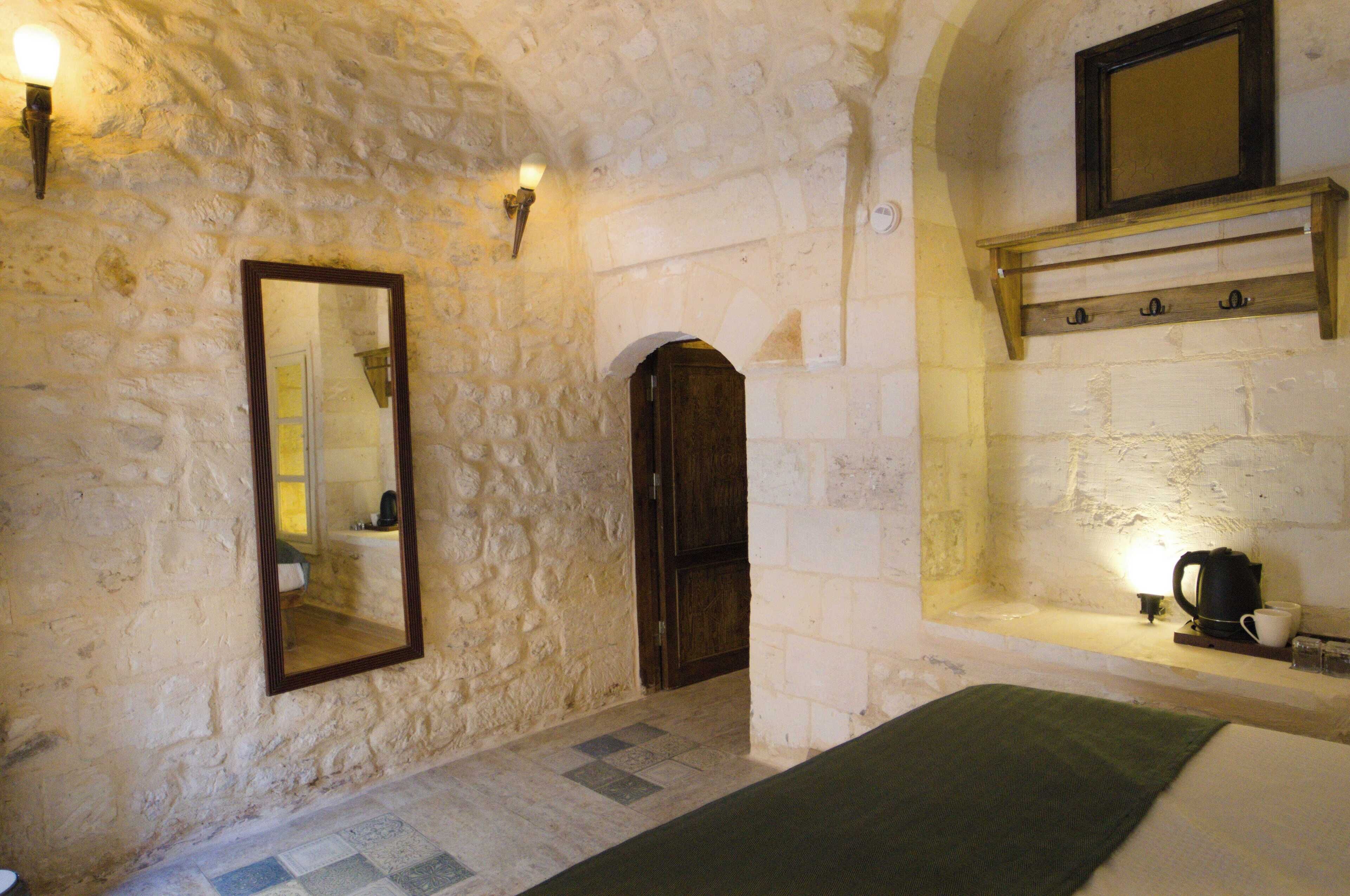 A stone-walled room with a large mirror, a wooden door, a shelf with hooks, and a small table with a kettle and cup.