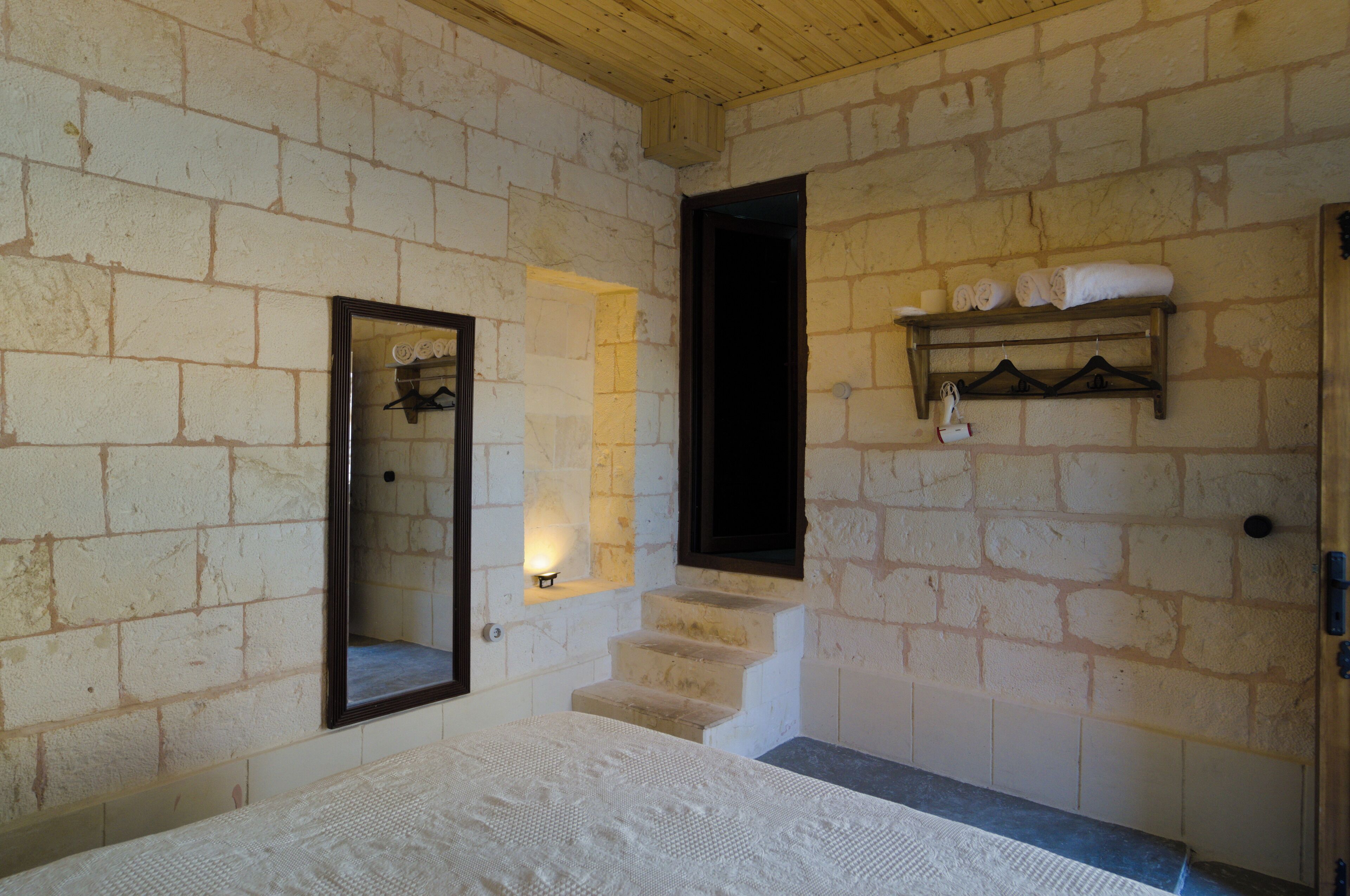 A bedroom with a bed, a wooden ceiling, a stone wall, a mirror, a shelf with towels, and a staircase.