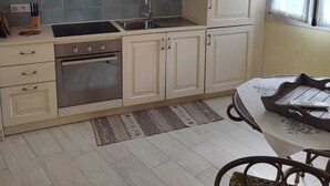A kitchen with white cabinets, a stainless steel sink, and a microwave. There is a window with curtains, a patterned rug, and a table with a chair.