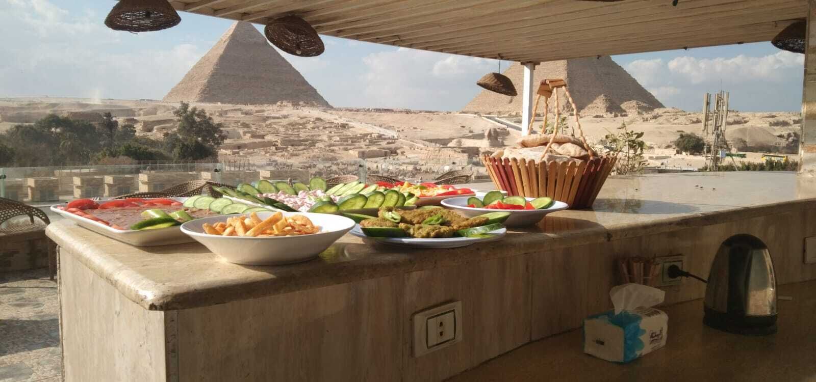 A buffet table with various dishes, a basket, and a kettle, set against a backdrop of pyramids.