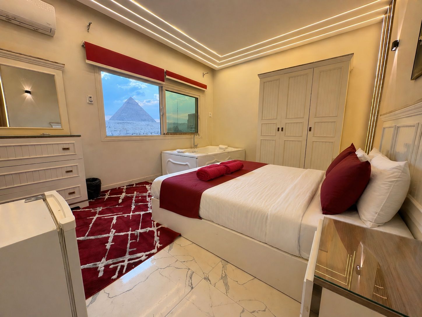 Une chambre d’hôtel comprenant un lit, une commode, une coiffeuse, une fenêtre donnant sur une pyramide et une armoire.
