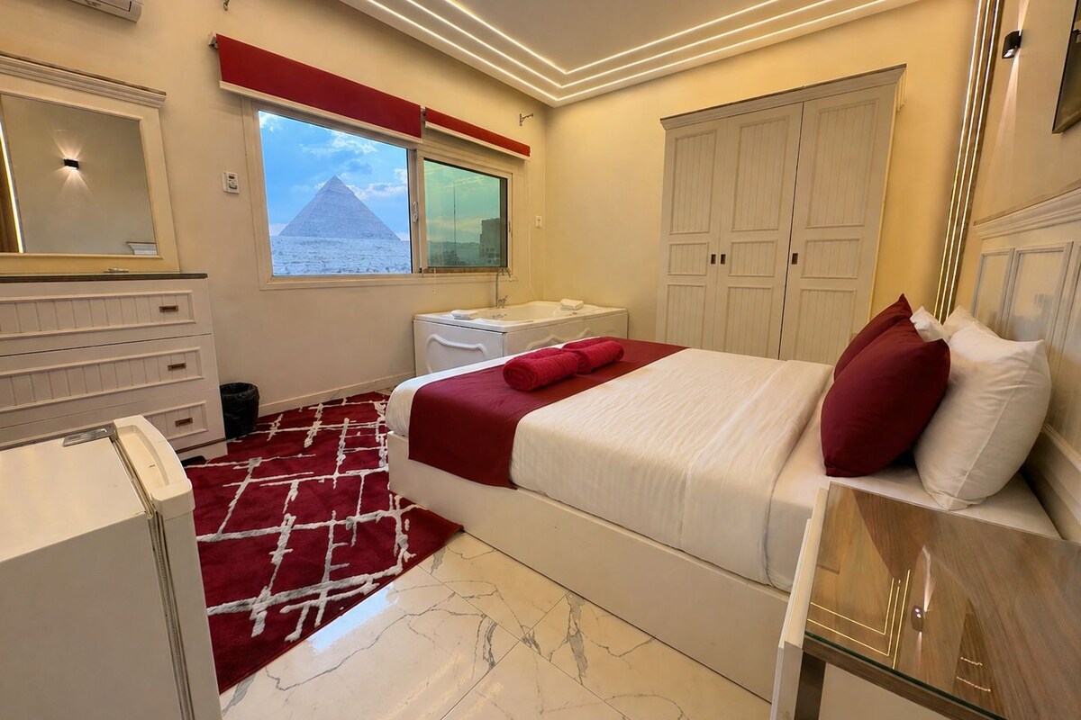 A hotel room with a bed, a dresser, a vanity, a window with a view of a pyramid, and a wardrobe.