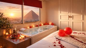 A room with a bathtub, a bed, a window with a view of a pyramid, and a potted plant.