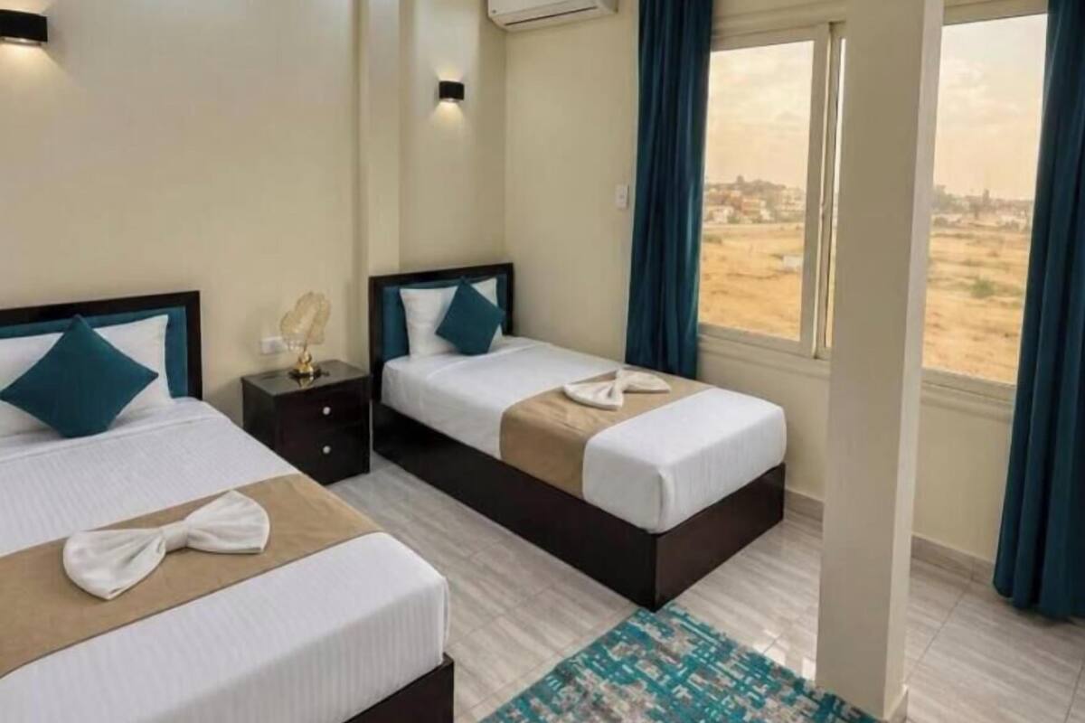 Una habitación de hotel con dos camas, una ventana con vistas, un equipo de aire acondicionado y una mesita de noche con una lámpara.