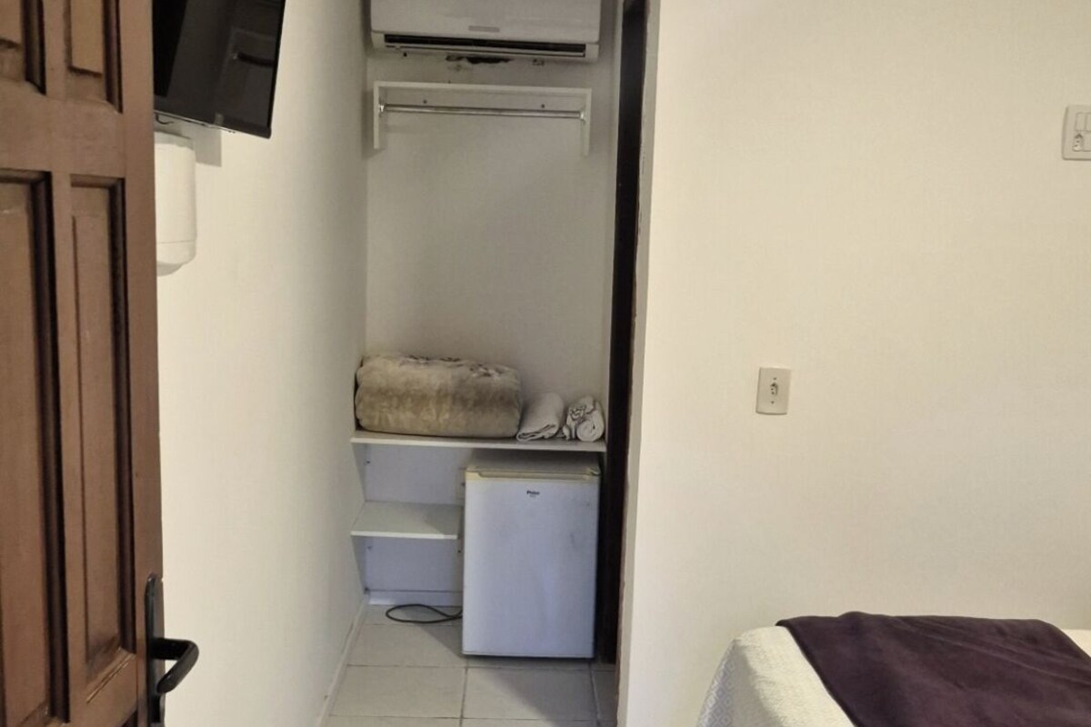 A narrow hallway with a bed on the right, a television on the left, and a small refrigerator in the center.