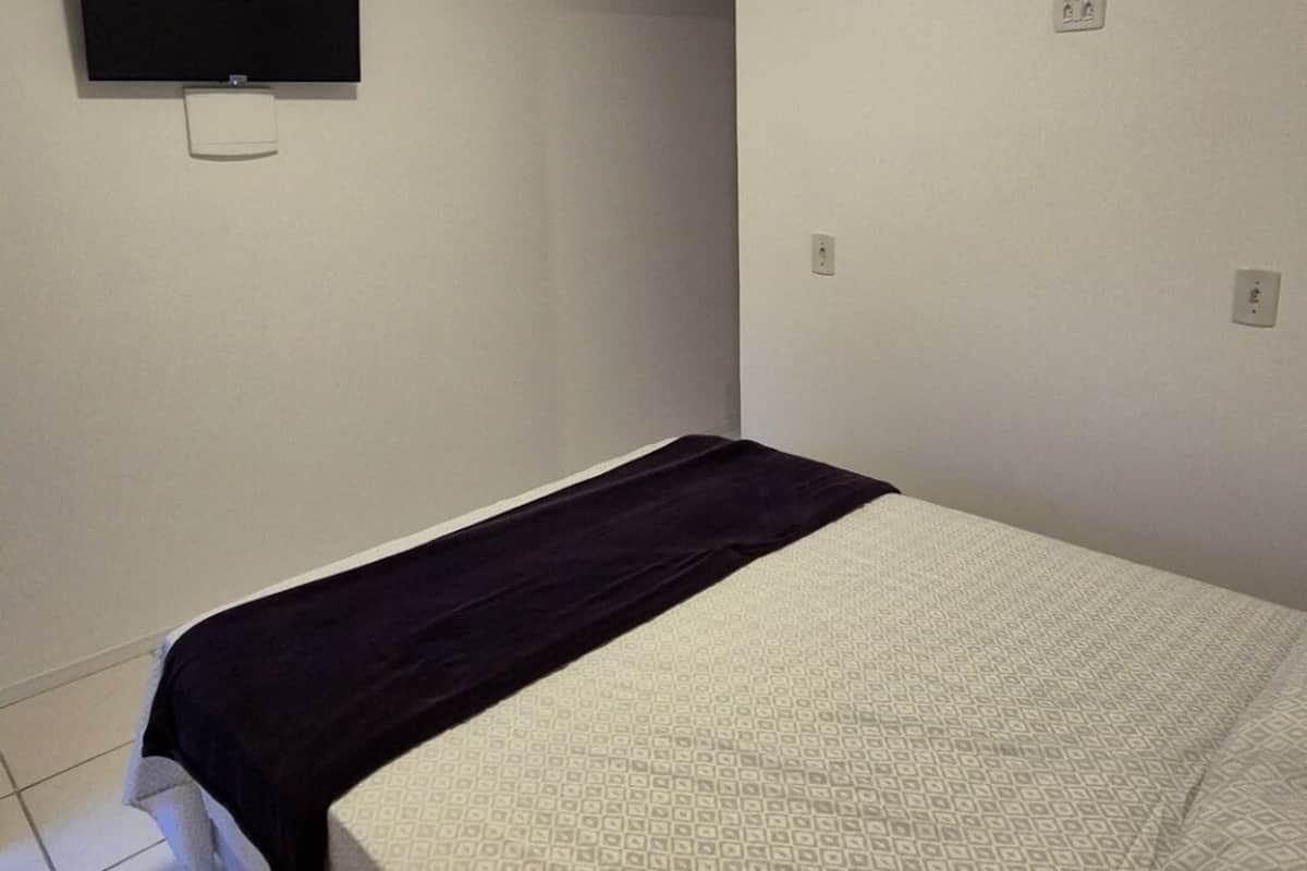 A small, sparsely furnished room with a bed, a flat-screen TV mounted on the wall, and a single power outlet.