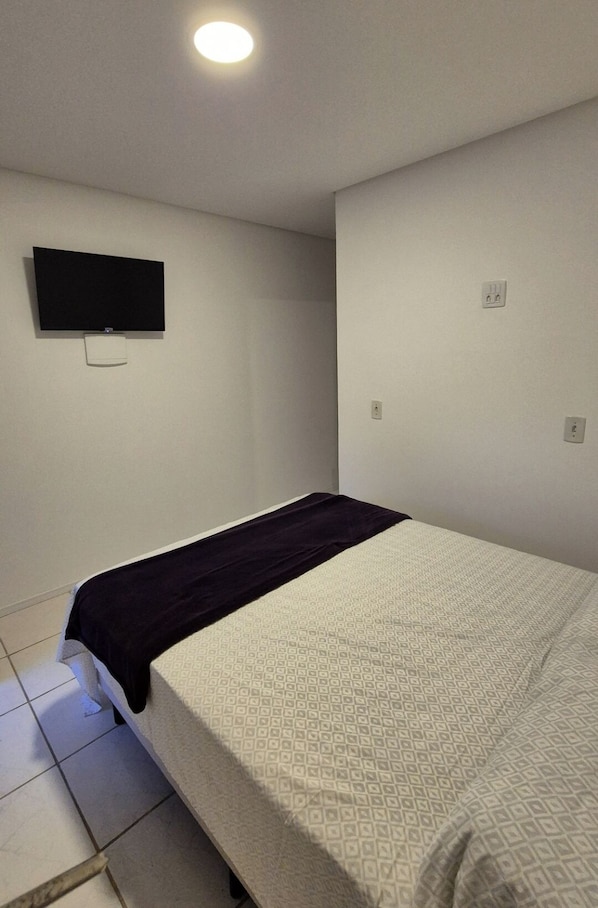 A small, sparsely furnished room with a bed, a flat-screen TV mounted on the wall, and a single power outlet. - Refugio Bela Ilha (Florianópolis)