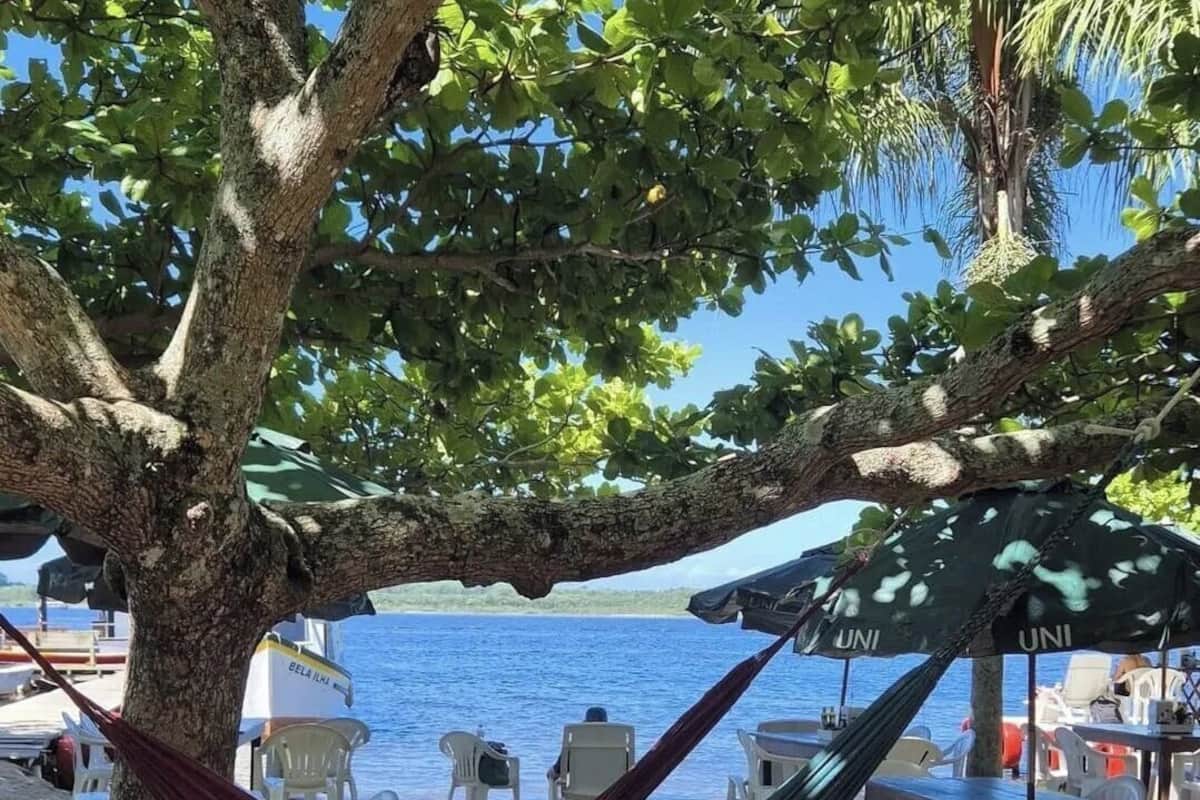 A hammock under a tree by a beach with a view of the sea, outdoor tables, and umbrellas.