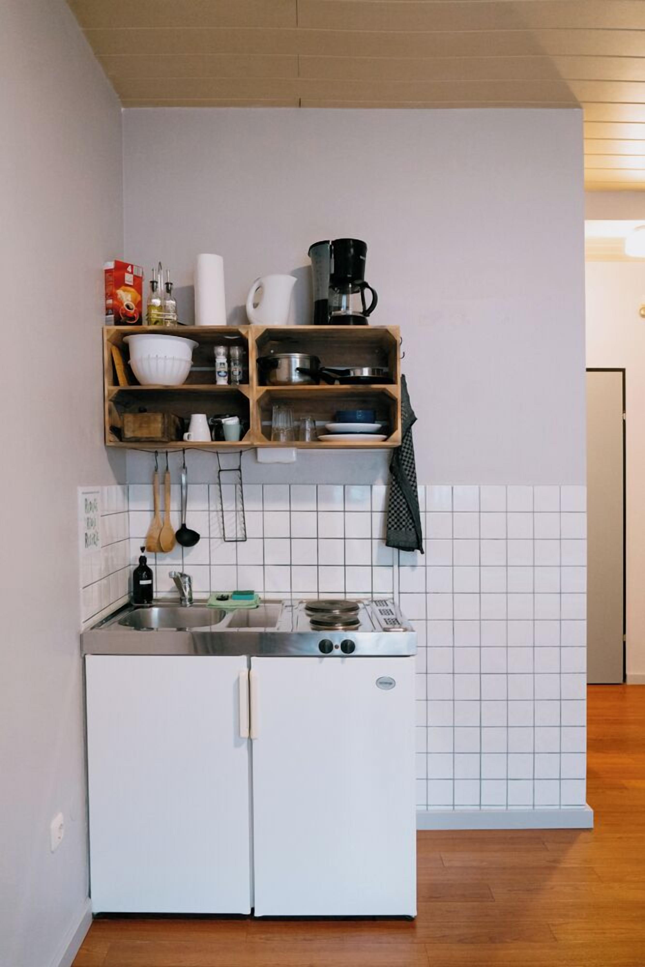 A compact kitchen with a white refrigerator, a small stove, and wooden shelves above the sink holding various kitchen items.