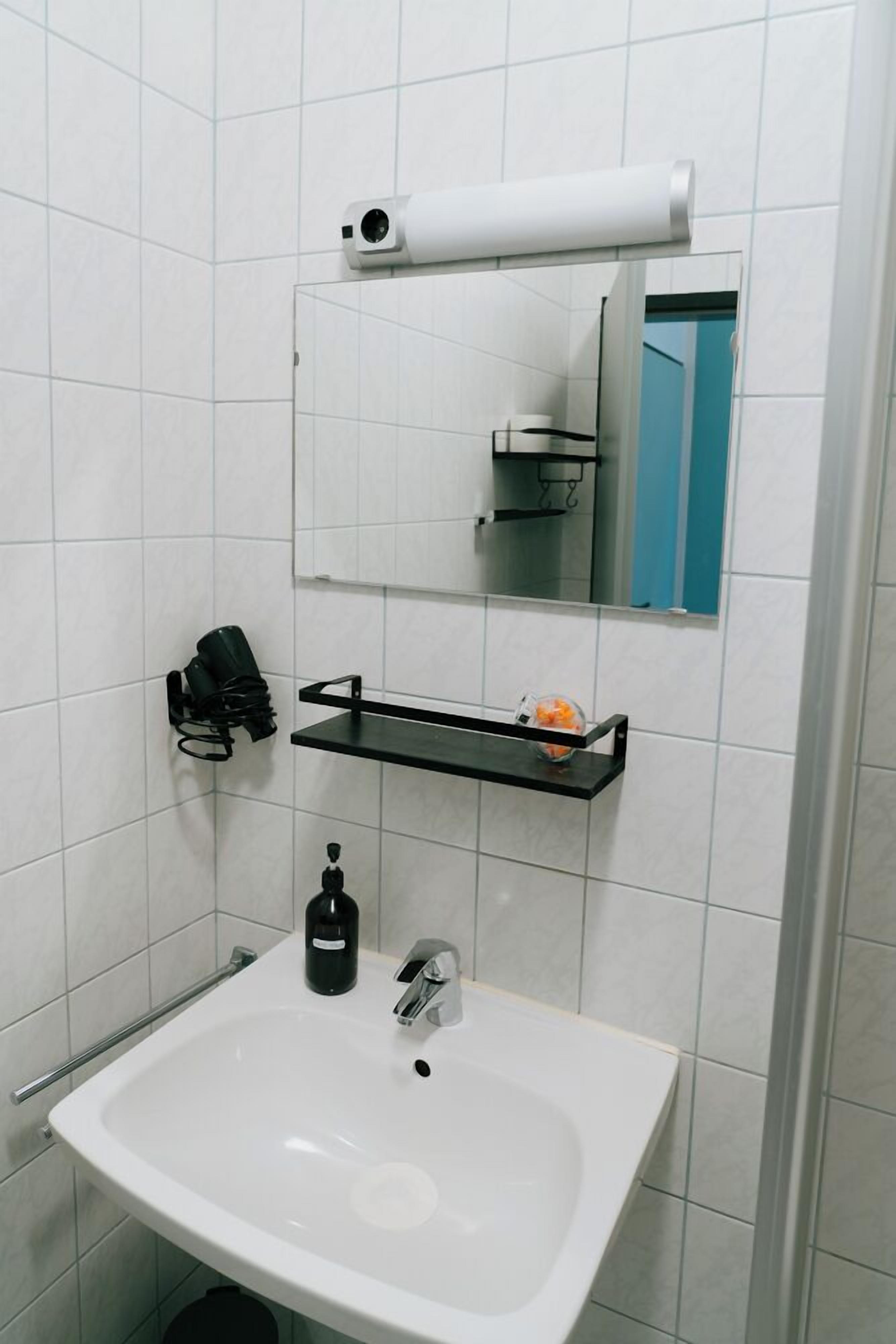 A bathroom with a white sink, a mirror, a towel dispenser, and a shelf with toiletries.