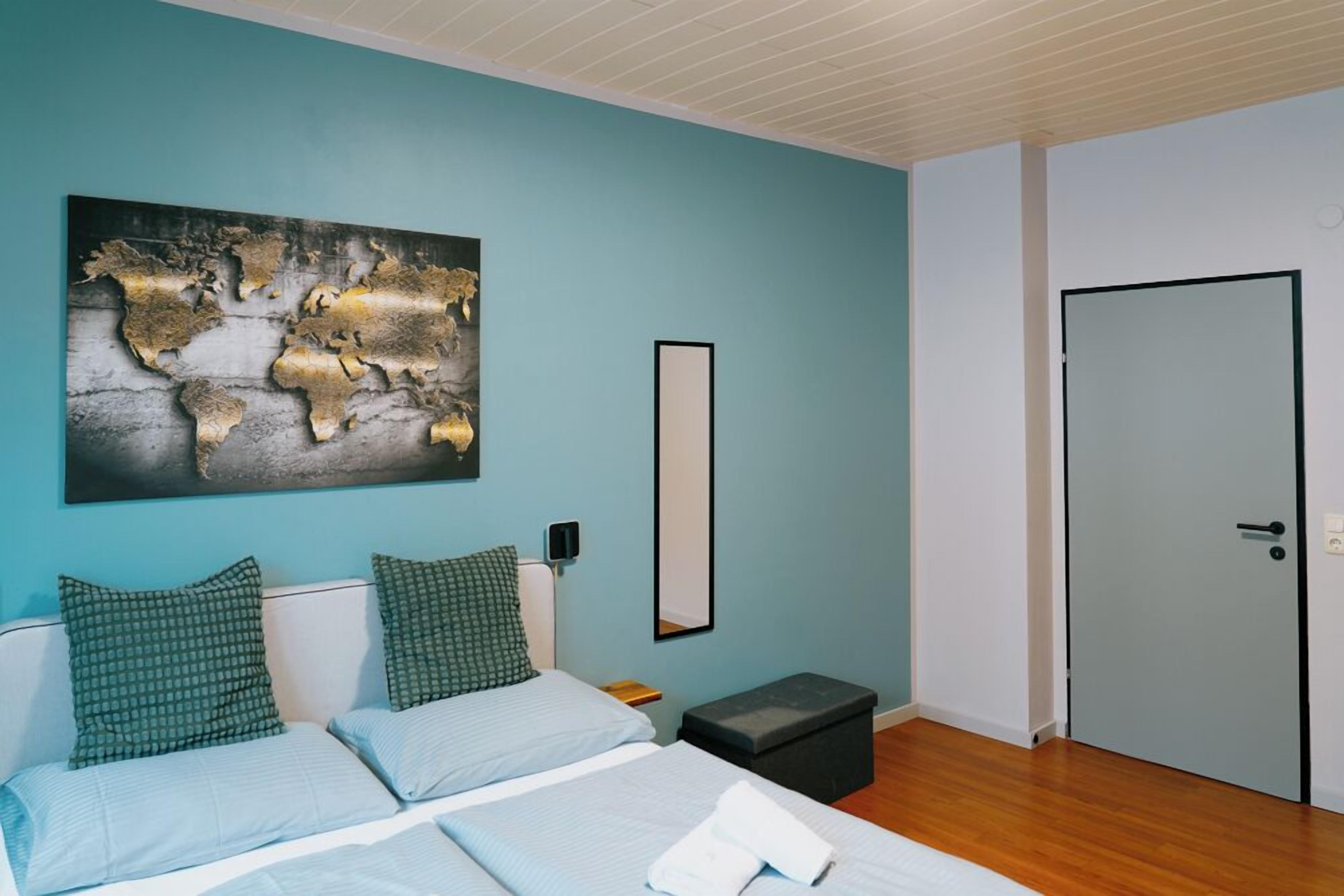 Un dormitorio con cama, dos almohadas, una mesita de noche, una pequeña mesa, una obra de arte con un mapa del mundo, un espejo y una puerta.