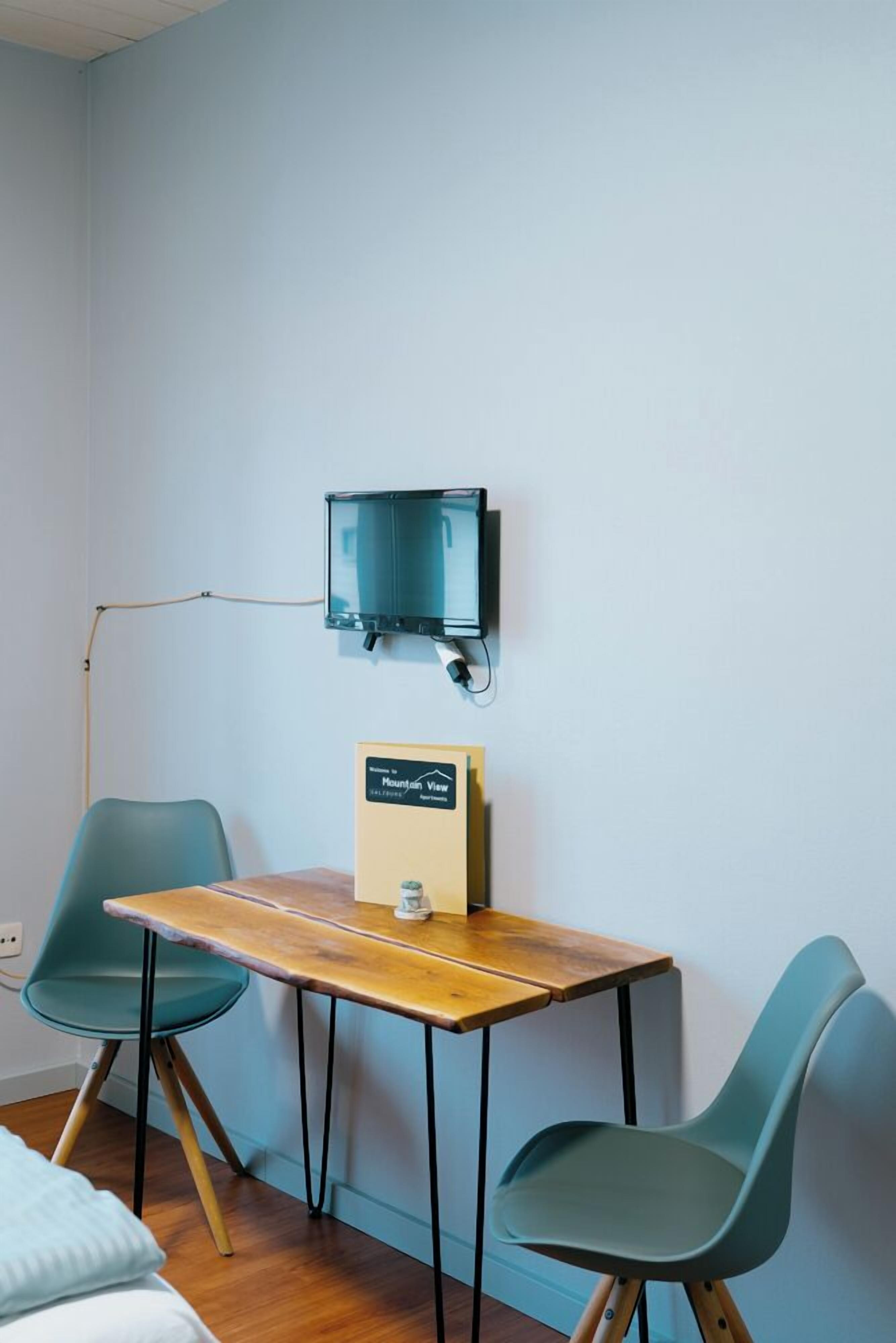 Ein kleines Zimmer mit einem Holztisch, zwei türkisfarbenen Stühlen, einem an der Wand befestigten Fernseher und einem Schild an der Wand.