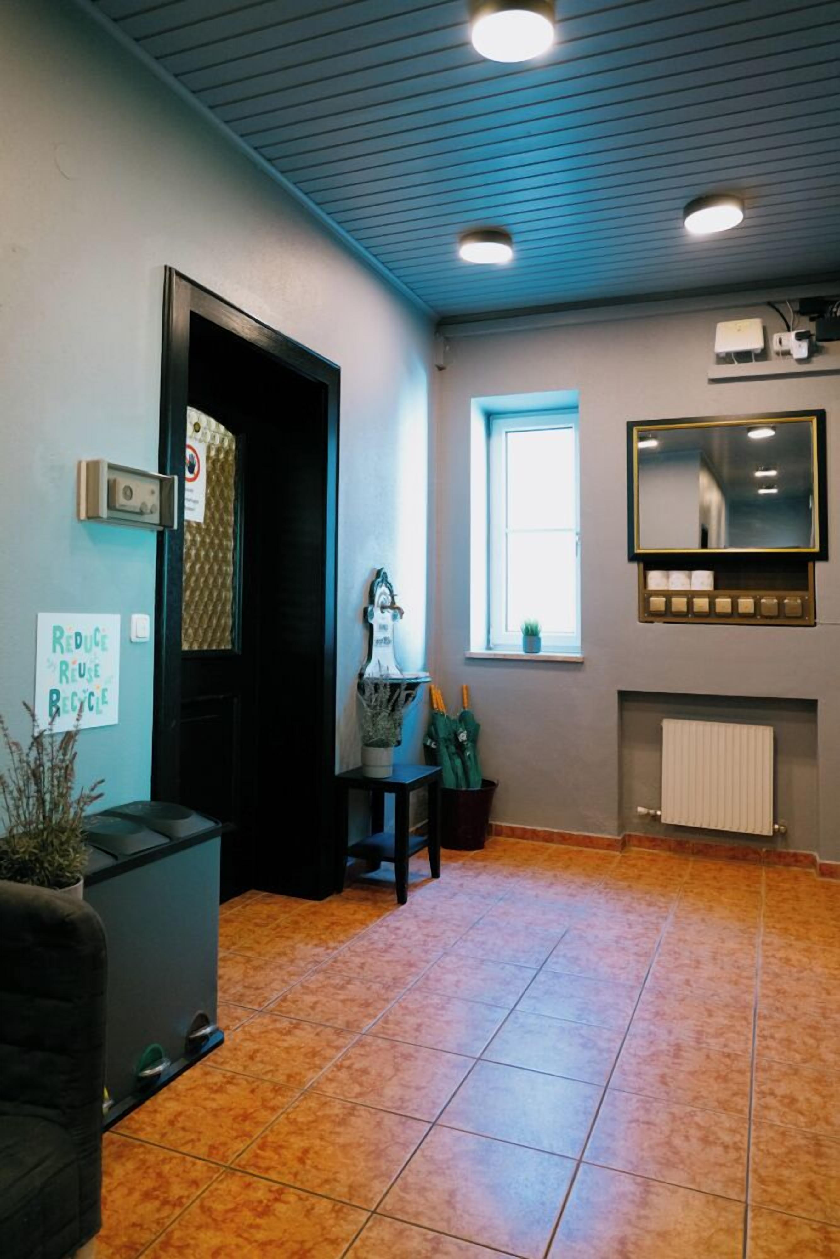 Una habitación con suelo de baldosas, una puerta negra, una ventana con persianas, un espejo sobre un radiador y un cartel en la pared.
