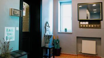 Una habitación con suelo de baldosas, una puerta negra, una ventana con persianas, un espejo sobre un radiador y un cartel en la pared.