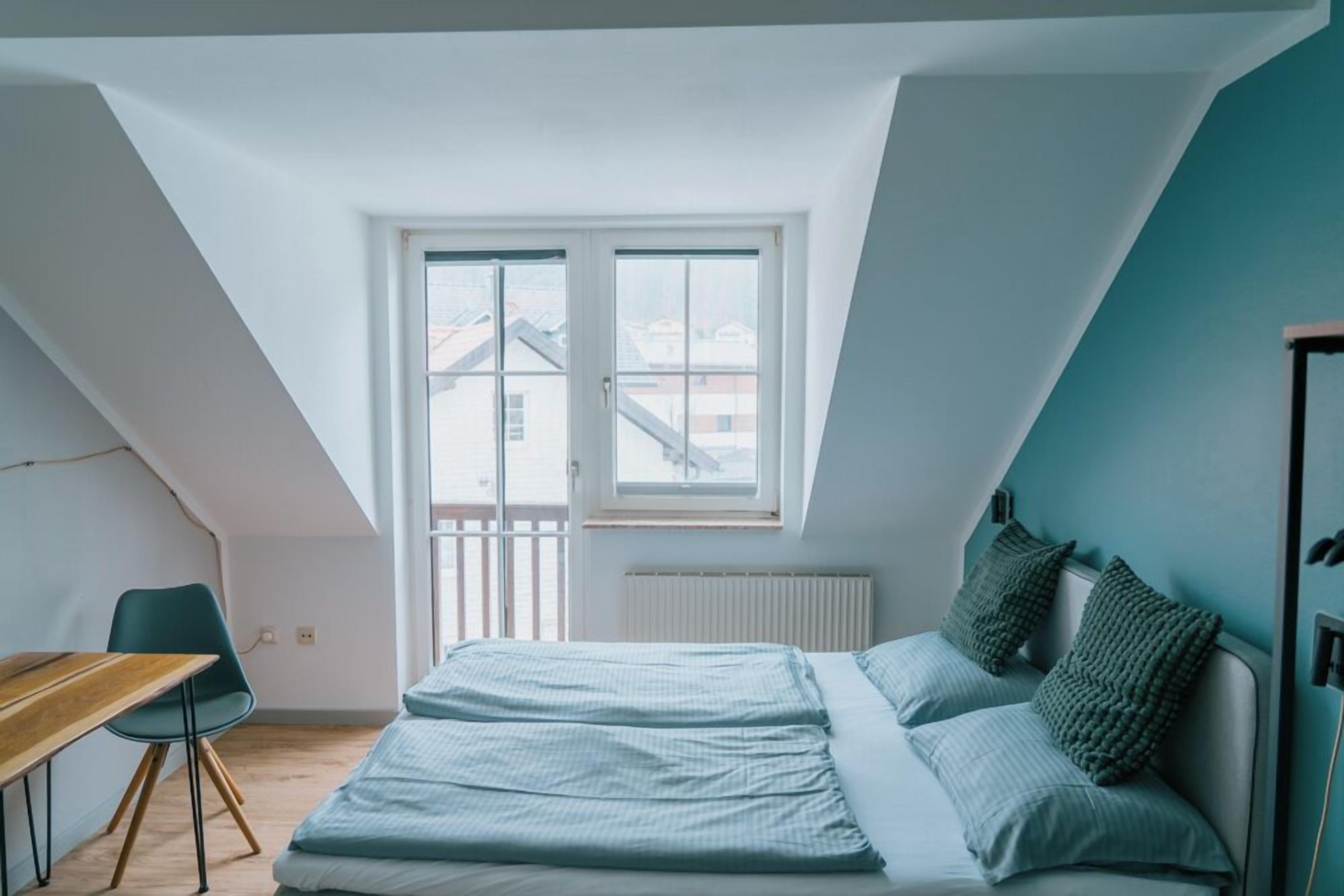 A bedroom with a sloped ceiling, a bed with green pillows, a desk with a chair, and a window with a view of buildings.