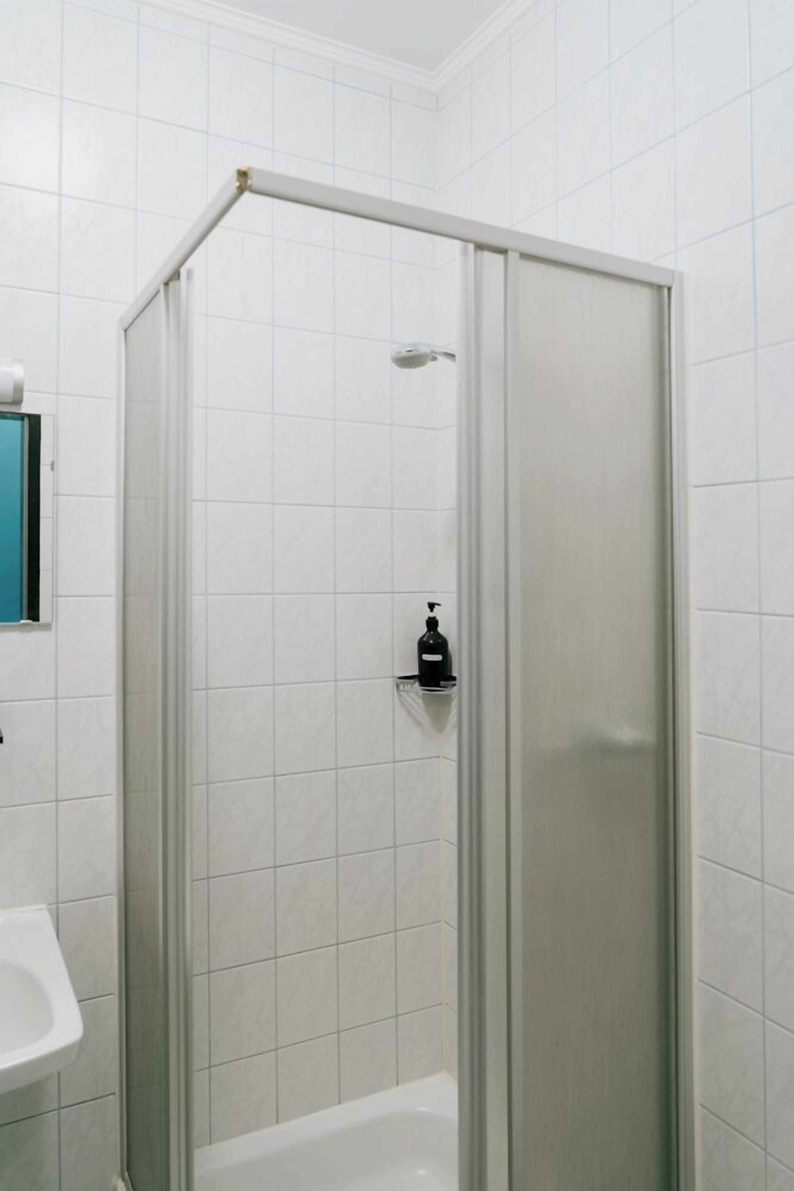 A shower with a glass door, white tiled walls, and a soap dispenser on the wall.