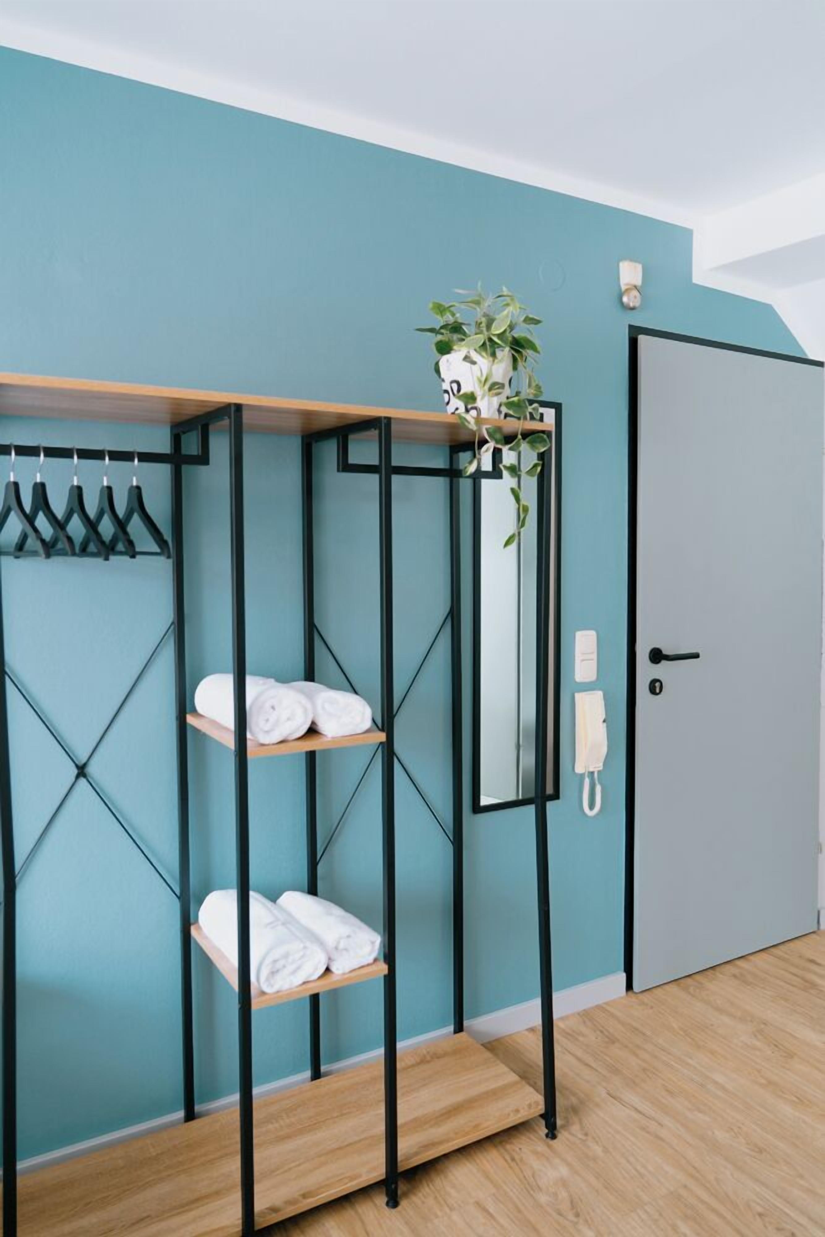 Una habitación con paredes verde azulado, piso de madera, un escurridor de metal con toallas, un espejo y una puerta.