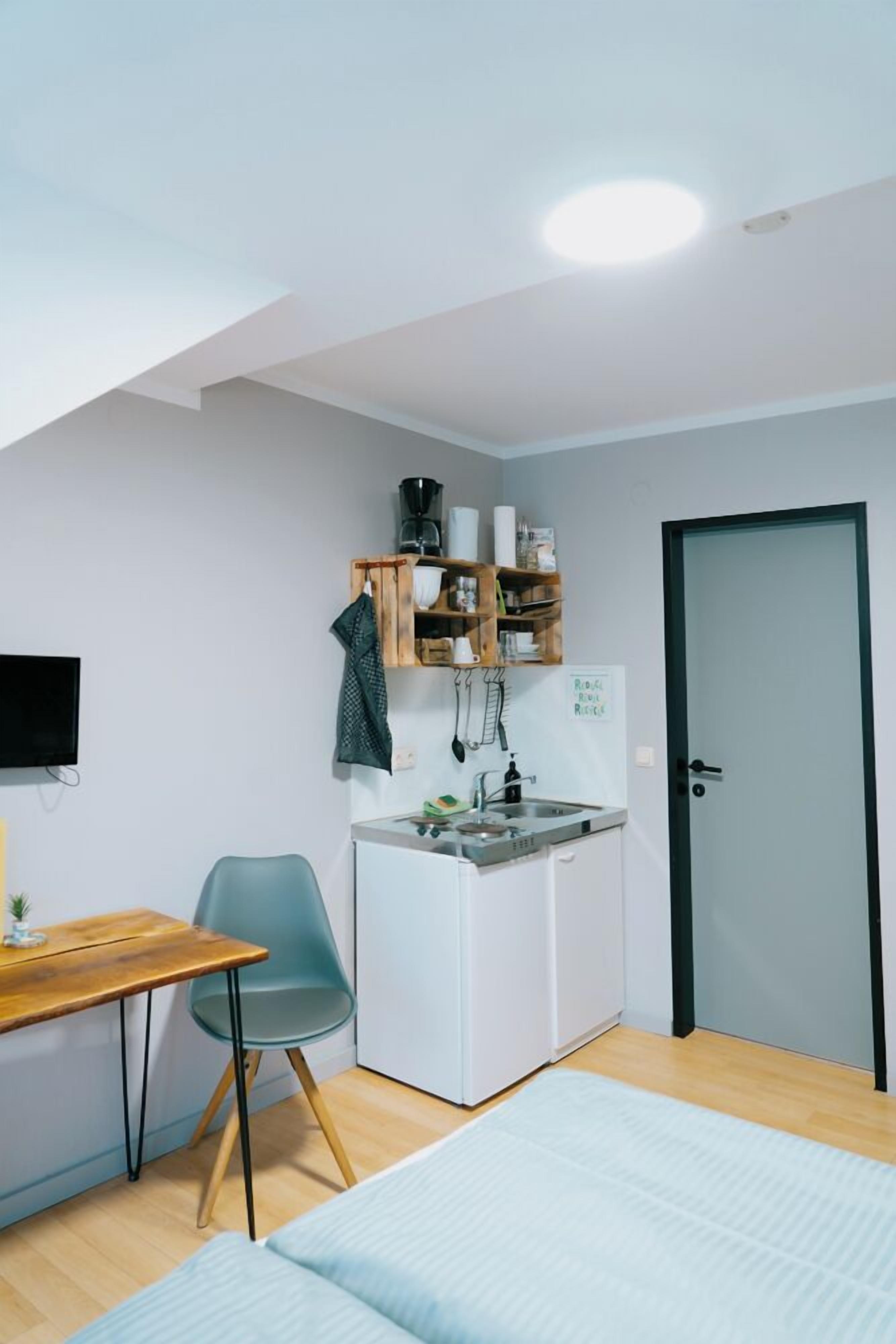 A compact kitchenette with a sink, stove, and storage shelves. A small dining table and chair are positioned near the window, and a bed is visible in the foreground.