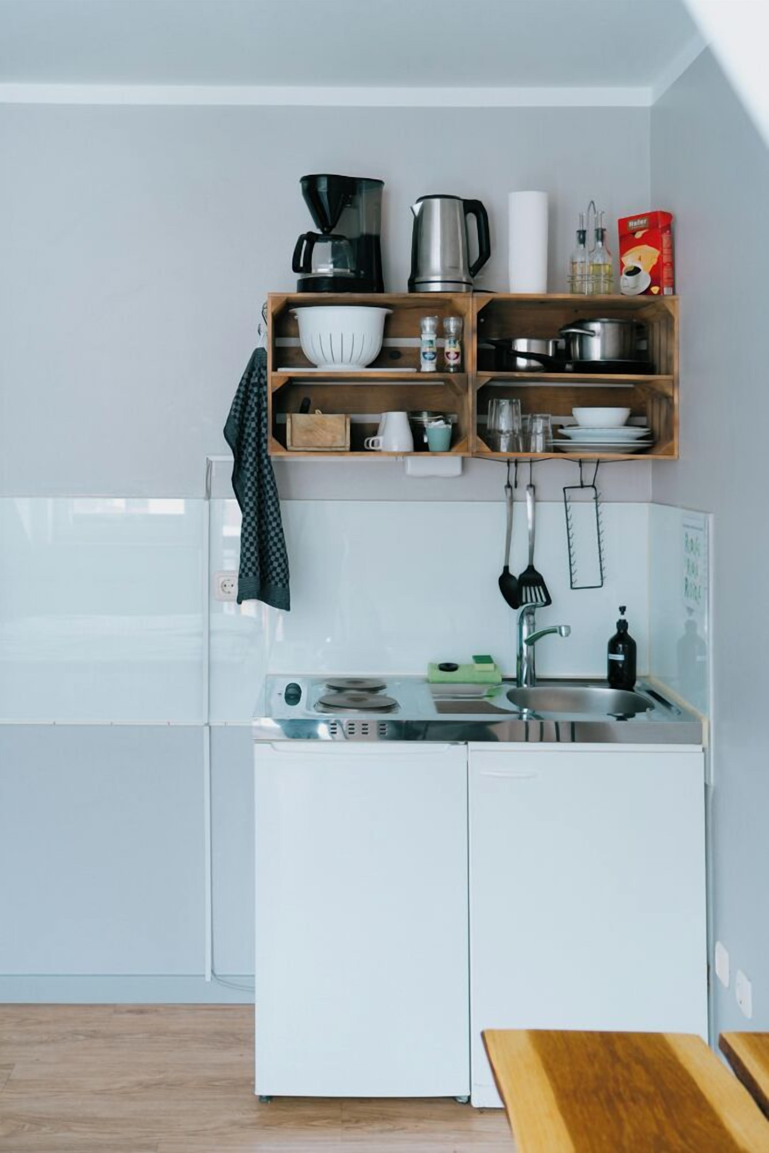 Eine kleine Küche mit einem weißen Kühlschrank, einem Spülbecken und Holzregalen, auf denen verschiedene Küchenutensilien stehen.