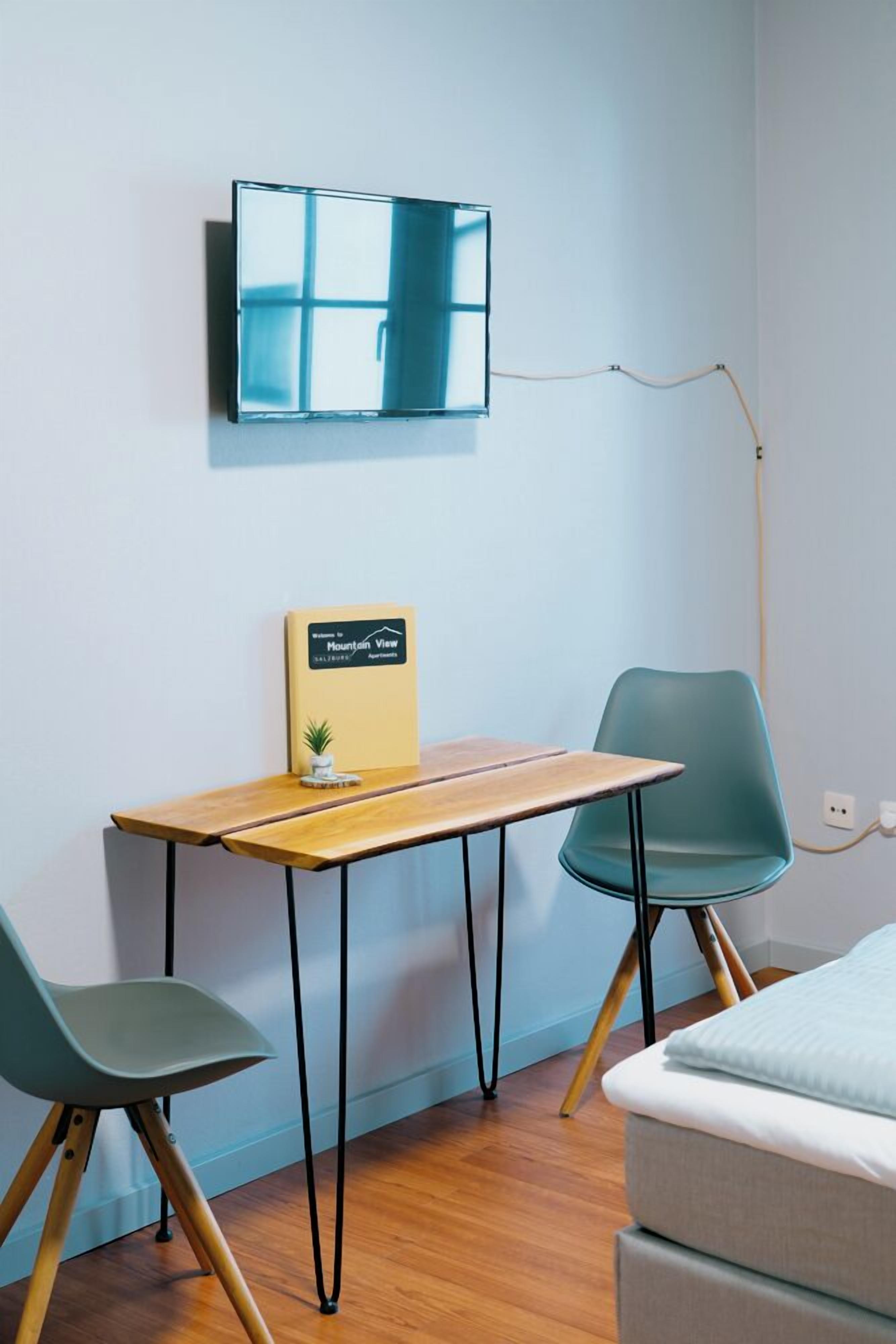 A room with a wooden desk, two chairs, a bed, a mounted TV, and a small plant.