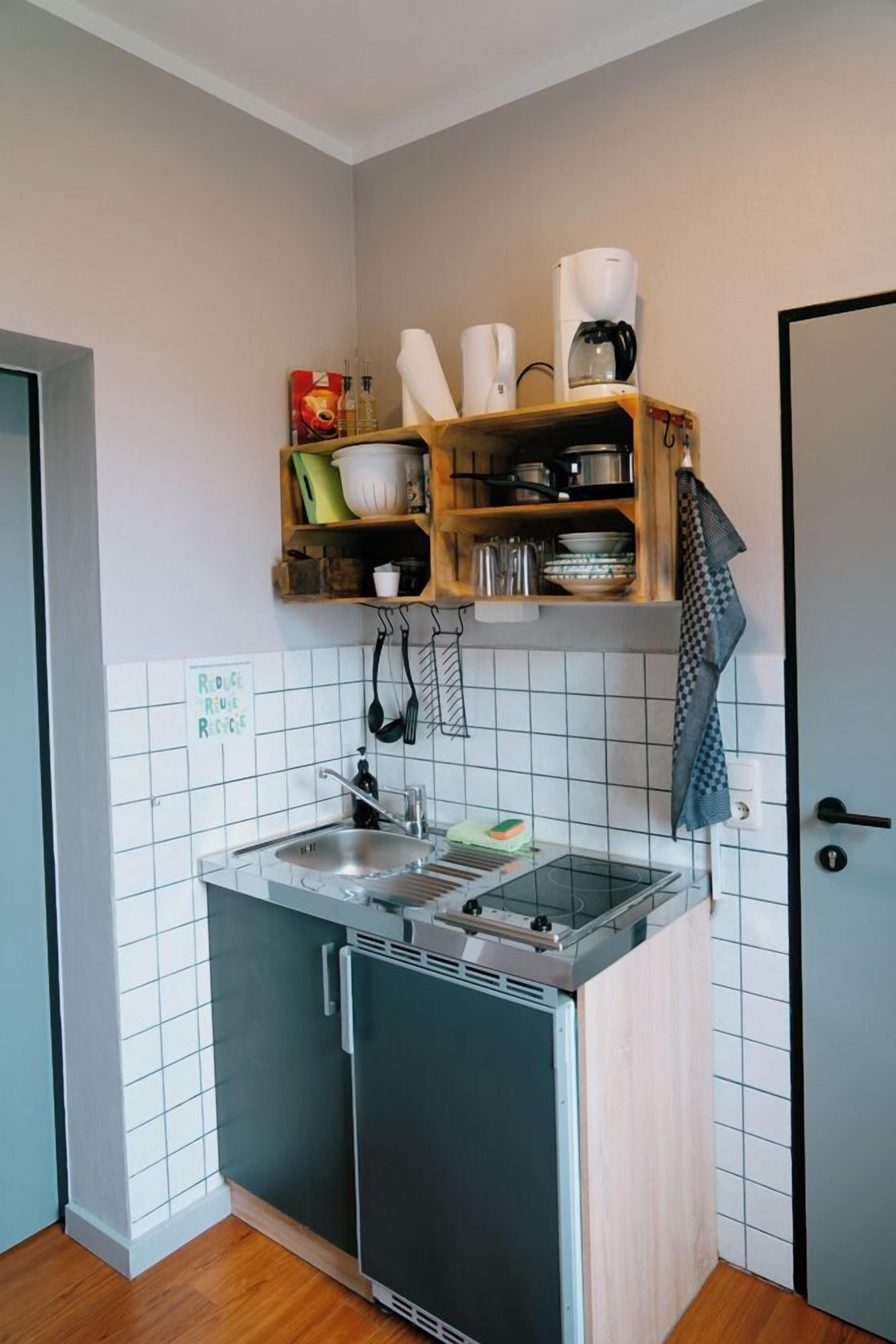 Una cucina compatta con piano cottura integrato, lavello e piccolo frigorifero. Sopra il piano cottura c'è una mensola in legno con stoviglie e un cartello appeso al muro.