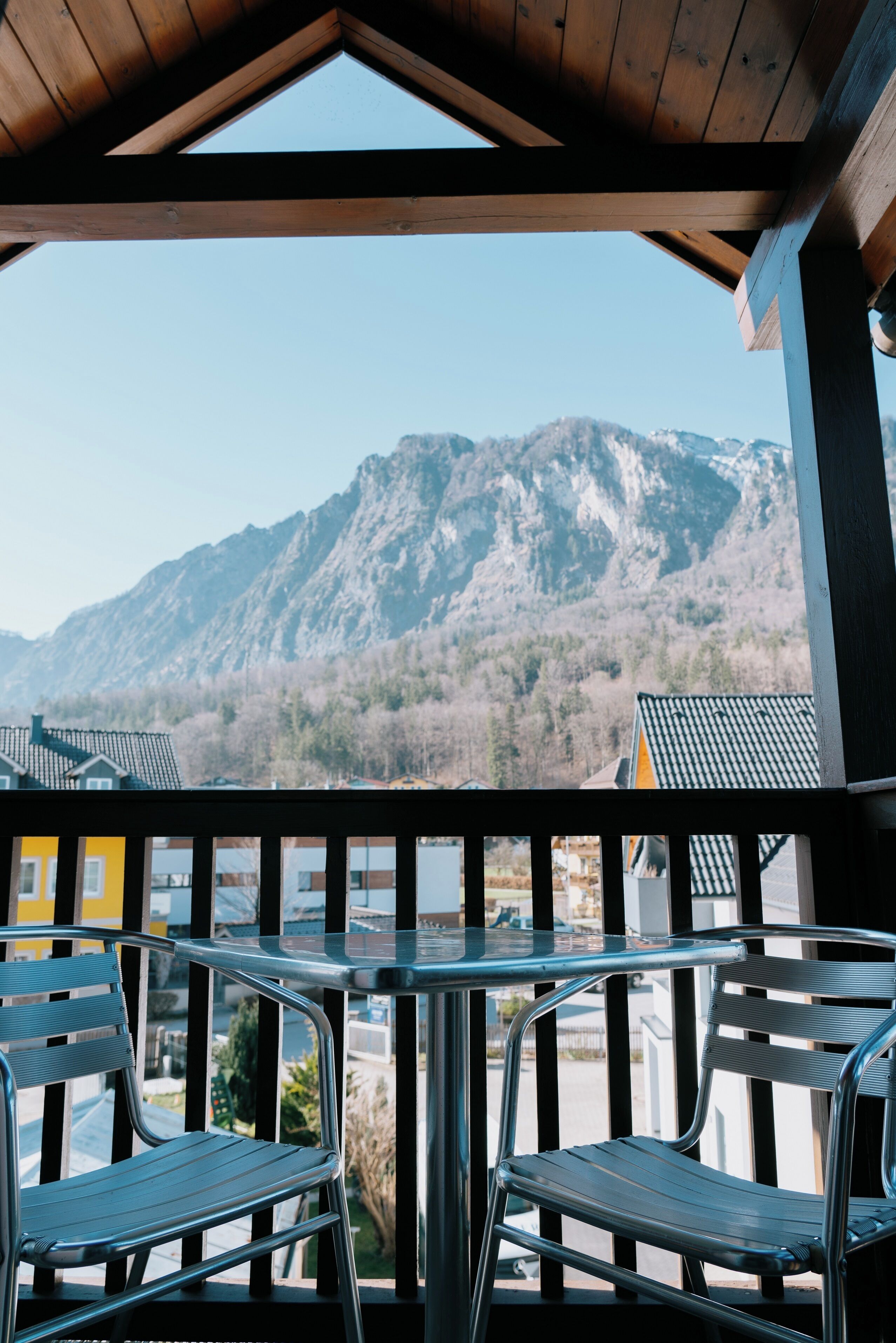 A balcony with metal chairs and a table, overlooking a mountainous landscape with buildings in the background.