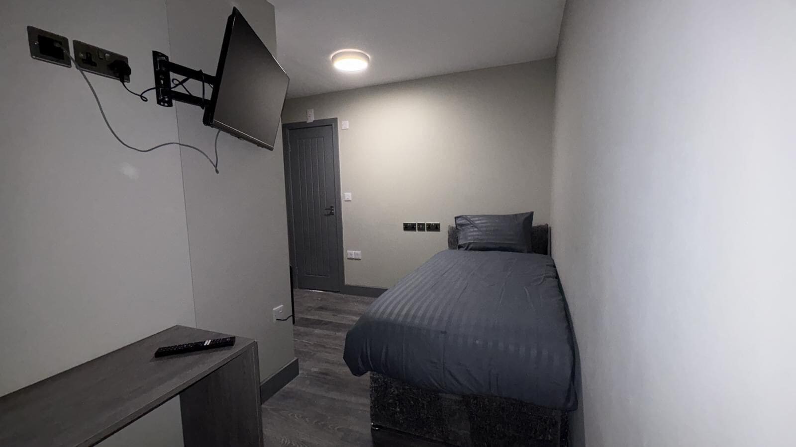 Une petite chambre à coucher moderne, équipée d’un lit, d’un téléviseur fixé au mur et d’un bureau avec une télécommande.