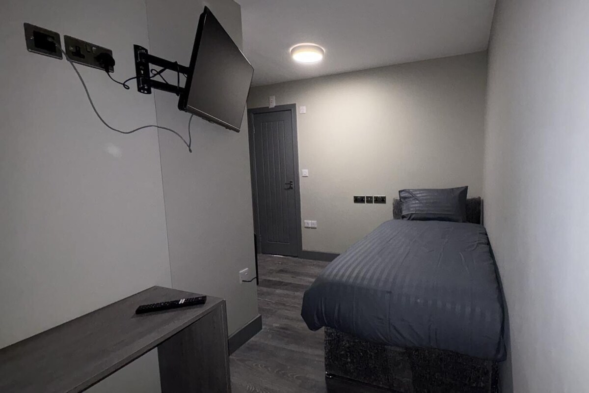 A small, modern bedroom with a bed, a TV mounted on the wall, and a desk with a remote control.