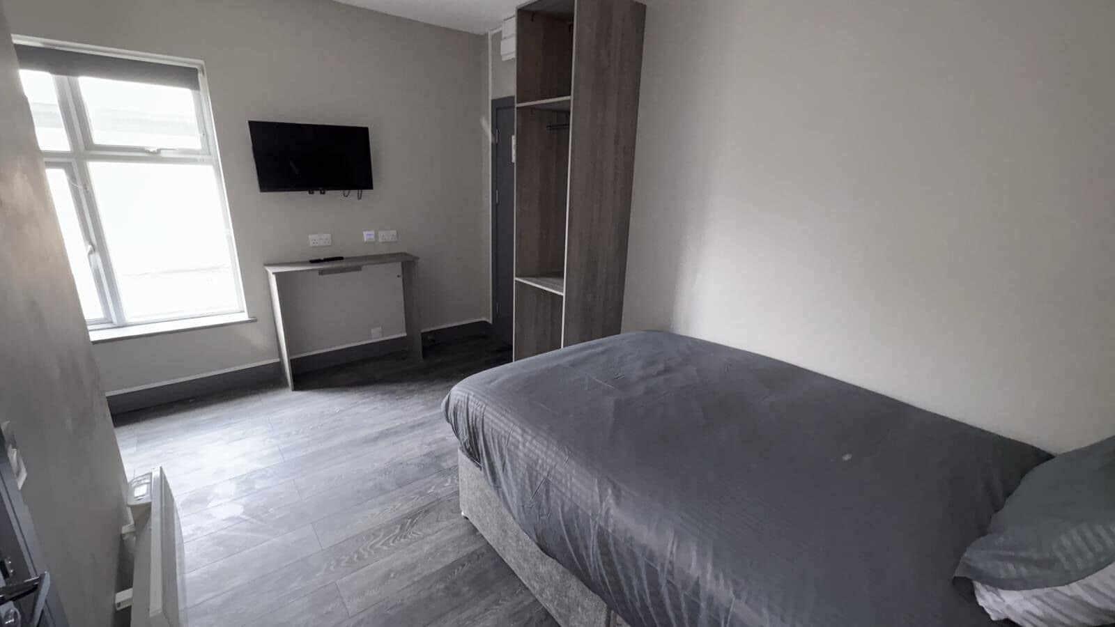 A bedroom with a bed, a TV mounted on the wall, a desk, and a wardrobe.