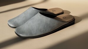 Une paire de chaussures grises, sans lacets, avec une bride noire et un logo sur la semelle, posée sur une surface beige.