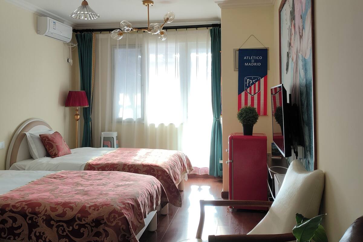 A hotel room with two beds, a desk, a chair, a small table, a potted plant, and a painting on the wall.