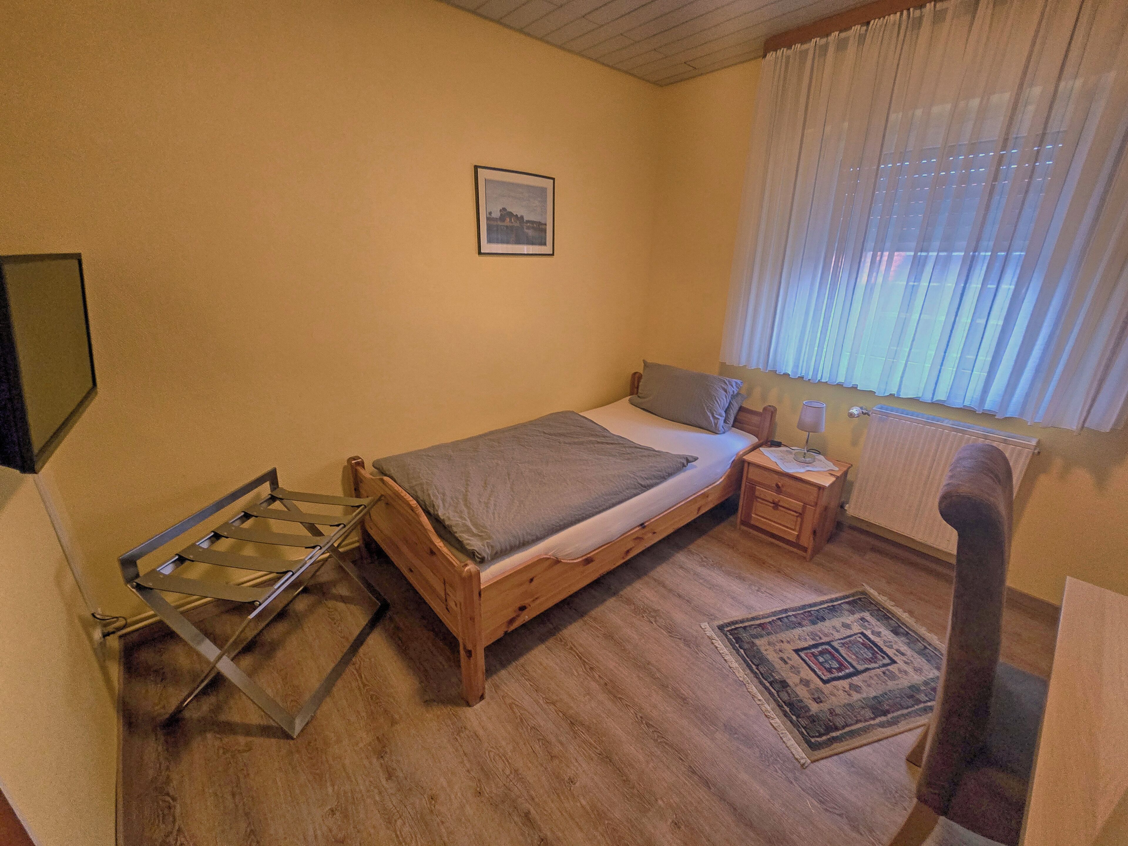 A bedroom with a wooden bed, a nightstand, a radiator, a window with curtains, and a picture on the wall.