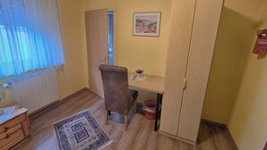 Een kleine kamer met een houten bureau, een stoel, een radiator, een houten kast, een lijst met een afbeelding aan de muur, en een raam met dunne gordijnen.