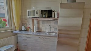 Een compacte keuken met een ingebouwde magnetron, een roestvrijstalen koelkast, een spoelbak en een raam met uitzicht op een gebouw en een plant.