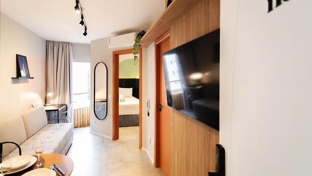 A modern hotel room with a large TV, a sofa, a dining table set, and a view of the city through the window. - Central Studium (São Paulo)