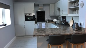 A modern kitchen with white cabinets, a built-in oven, a kettle, and a coffee maker. There are two bar stools and a window with a view of the outside. - Willow Cottage at Sound Healing Centre (Cardigan)
