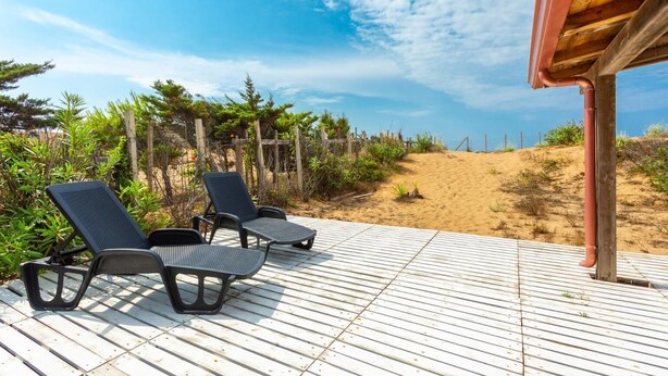 Une terrasse en bois avec deux chaises longues donnant sur une plage de sable et un ciel dégagé.
