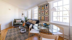 Een moderne woonkamer met een glazen tafel, witte stoelen, een groene bank en een kamerbreed tapijt met een patroon.
