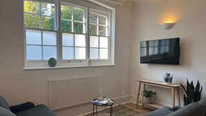 Un salon avec une grande fenêtre, un téléviseur à écran plat, un radiateur, une petite table avec des magazines et une plante en pot.