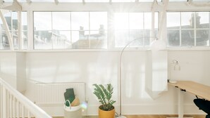 Um quarto iluminado com grandes janelas, uma escrivaninha de madeira, um abajur e um vaso com planta.
