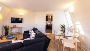 Ein modernes Wohnzimmer mit einem schwarzen Sofa, einem Glastisch und einer Küche im Hintergrund.