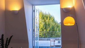 Ein modernes Wohnzimmer mit einem dunklen Sofa, gemusterten Kissen und einer Stehlampe. Ein großes Fenster bietet Blick auf einen Balkon mit Geländer und Bepflanzung.