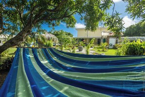 Relax in a shaded hammock suspended between two trees, surrounded by lush plants and a well-manicured lawn.