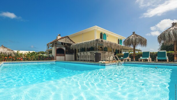 A clear blue pool with lounge chairs and umbrellas, a building with a thatched roof, and a clear sky.