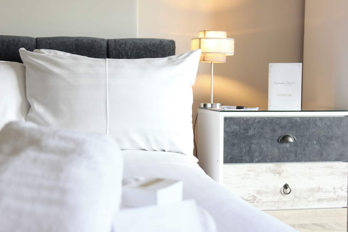 Una cama bien hecha con ropa blanca, un cabecero gris, una lámpara de noche y una mesita de madera con un cajón.