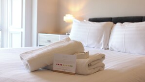 Een keurig opgemaakt bed met witte lakens en een gevouwen handdoek, een nachtkastje met een lamp, en een raam met natuurlijk licht.