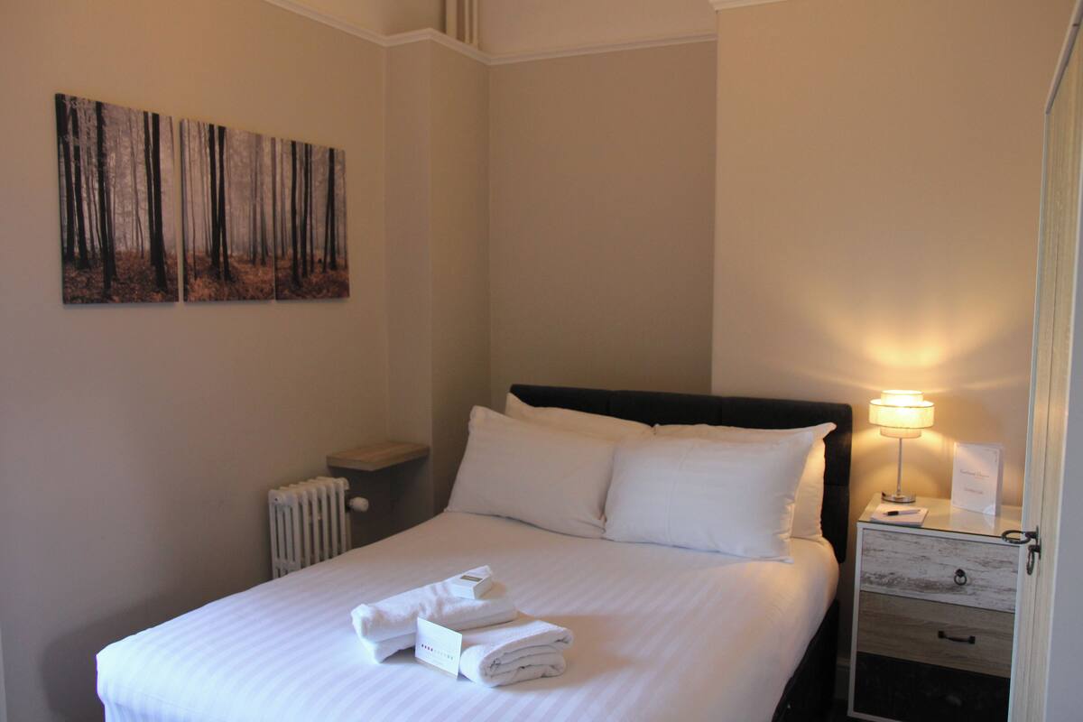 Una cama bien hecha con ropa y toallas blancas, una lámpara de noche, una mesita de noche con cajón y un cuadro de un bosque en la pared.