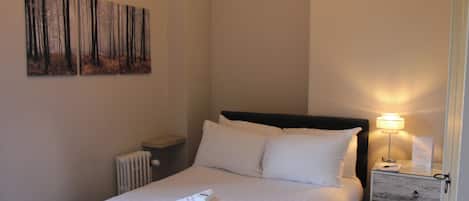 Una cama bien hecha con ropa y toallas blancas, una lámpara de noche, una mesita de noche con cajón y un cuadro de un bosque en la pared.